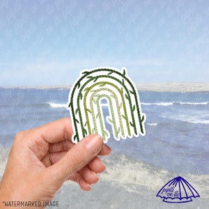 May include: A sticker featuring a green and white rainbow design. The rainbow is composed of rope-like textures and leafy vines. The sticker is held in a hand against a backdrop of a beach and ocean.