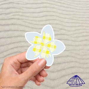 May include: A light blue flower sticker with a yellow plaid pattern. The sticker is being held in a hand.
