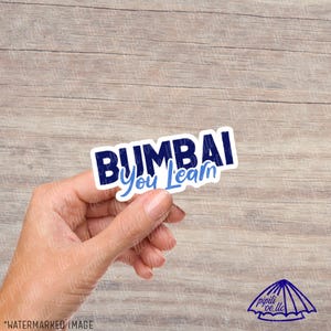 May include: A white sticker with the text "BUMBAI You Learn" in navy blue and light blue. The sticker is held by a hand against a wooden background. The sticker has a white border.