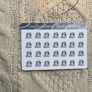 May include: A sticker sheet with numerous small illustrations of sewing machines in light blue and grey. The text "CozyBookSleeve - Sewing Machine" is printed at the top. The sticker sheet is on a cream-coloured knitted surface.