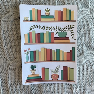 May include: A sticker sheet with four shelves of books, plants, and a cup of coffee. The shelves are white with brown trim. The books are various colors, including red, green, yellow, and brown. The plants are green and brown. The cup of coffee is white with a red heart.