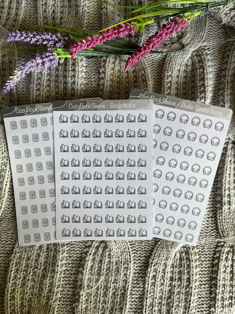 Op de afbeelding: Drie vellen met stickers met zwarte en witte pictogrammen. Het eerste vel heeft een raster met kleine vierkanten. Het tweede vel heeft een raster met open boekpictogrammen. Het derde vel heeft een raster met koptelefoonpictogrammen. De tekst op de vellen luidt "Cozy Book Sleeve - Book Icons" en "Cozy Book Sleeve - Audio Books Icons".