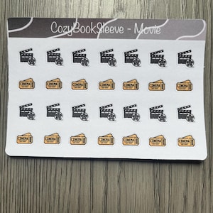 May include: A sticker sheet with film-related designs. The stickers show black clapperboards with film reels and yellow cinema tickets with the word "CINEMA". The text "CozyBookSleeve - Movie" is printed at the top of the sheet.