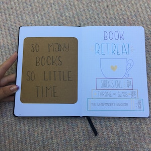 May include: A black notebook open to two pages. The left page has a brown insert with the text "SO MANY BOOKS SO LITTLE TIME". The right page says "BOOK RETREAT" with a teacup graphic and book titles: "SIREN'S CALL", "THRONE OF GLASS", and "THE WATCHMAKER'S DAUGHTER".