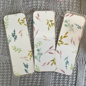 Può includere: Tre segnalibri rettangolari con angoli arrotondati. Ogni segnalibro presenta uno sfondo bianco con un motivo ad acquerello di foglie e ramoscelli verdi, rosa e color oliva. I segnalibri sono protetti da un rivestimento di plastica trasparente.