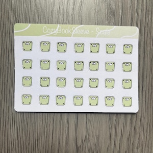 Peut inclure: Une planche d'autocollants avec de nombreuses petites illustrations de balances vert clair. Chaque balance est carrée avec un dessus arrondi et un petit cadran. Le texte "Cozy Book Sleeve - Seate" est en haut de la feuille.