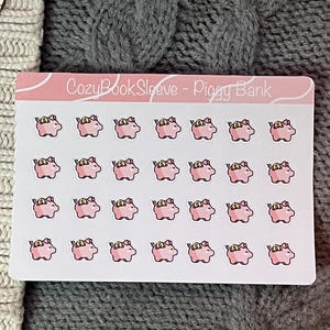 May include: A sheet of stickers featuring pink piggy bank illustrations with gold coins. The stickers are on a white background with the text "CozyBookSleeve - Piggy Bank" in pink. The background is a grey knitted fabric.