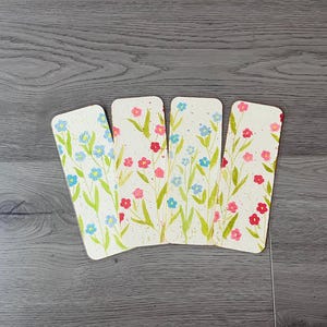 May include: Four rectangular bookmarks with rounded corners, each featuring a watercolour floral design. The flowers are in shades of blue and red, with green stems and leaves, set against a speckled off-white background.
