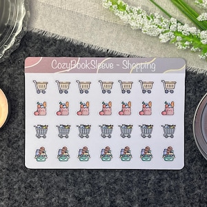 May include: A sheet of stickers featuring various shopping cart designs. The stickers are white with a pink, peach, and green color scheme. The text "CozyBookSleeve - Shopping" is at the top of the sheet.
