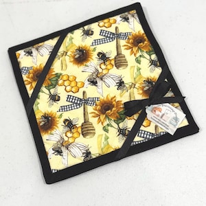 May include: Square quilted pot holder with a yellow background featuring sunflowers, bees, and honey dippers. The pot holder is trimmed with black binding and has a black ribbon accent. A small tag is attached to the ribbon.