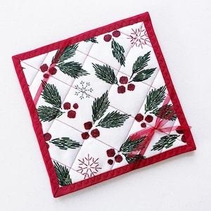 May include: Square quilted Christmas potholder with a white background. The potholder features a pattern of green holly leaves, red berries, and snowflakes. It has a red border and a red ribbon tied in a bow.