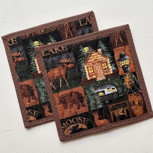 Op de afbeelding: Twee vierkante, gewatteerde onderzetters met een bruine rand. Elke onderzetter heeft een patchworkontwerp met afbeeldingen van hutten, elanden en meergezichten. De onderzetters hebben de woorden "CABINS", "LAKE", "MOOSE" en "LAKEVIEW" erop gedrukt.