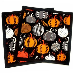 Könnte beinhalten: Set aus drei schwarzen, gesteppten Topflappen mit Halloween-Motiv. Die Topflappen zeigen orange, weiße und graue Kürbisse auf schwarzem Hintergrund.