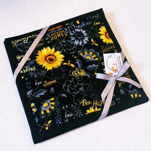 May include: Black quilted square with a sunflower and bee pattern. The design includes yellow sunflowers, grey and yellow floral accents, and the words "Sunshine", "Happy Bees", and "Bee Sweet". A grey ribbon is tied around the square.