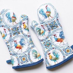 May include: A pair of white quilted oven mitts with a blue and white rooster pattern. The mitts feature a repeating design of roosters in medallions, with blue and yellow accents. The edges are trimmed with a blue binding and a blue loop.