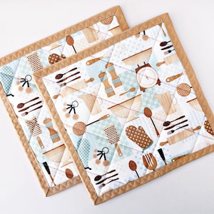Puede incluir: Dos salvamanteles cuadrados acolchados con un fondo azul claro y blanco con utensilios de cocina marrones y beige. Los bordes están ribeteados con una tela marrón. Los salvamanteles están acolchados con un patrón de diamantes.
