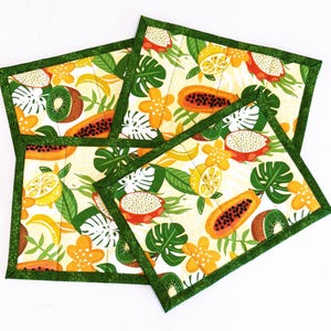 May include: Four green-trimmed placemats with a tropical fruit design. The placemats feature a white background with illustrations of papaya, kiwi, lemons, dragon fruit, and monstera leaves.