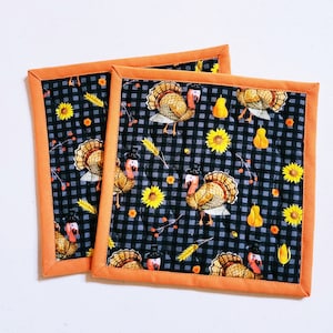 May include: Two square quilted pot holders. The fabric features a black and gray checkered pattern with illustrations of turkeys, sunflowers, and pumpkins. The pot holders have an orange border.
