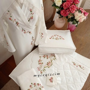 May include: White embroidered robe, pillow, and prayer rug with pink rose designs. The set includes a small towel with a rose design and a tassel. The text "myceyizcim" is visible on the rug.