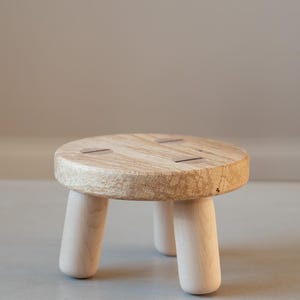May include: A small, round wooden stool with three short, rounded legs. The top is made of light-coloured wood with a textured surface and four small, dark-coloured inlays. The legs are a lighter shade of wood. The stool is approximately 25 cm in diameter.