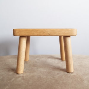 Puede incluir: Un pequeño taburete de madera con cuatro patas. El taburete está hecho de madera marrón claro y tiene una parte superior rectangular.