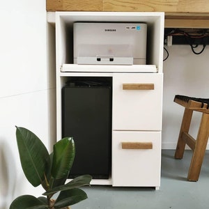 May include: A white storage cabinet with two drawers and a top shelf. A Samsung printer sits on the top shelf. The drawers have wooden handles. A black rectangular object is in the cabinet. A potted plant with large green leaves is in the foreground.
