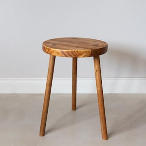 Pode incluir: Um pequeno banco de madeira de três pernas com um assento redondo. O banco é feito de madeira castanha clara e tem um acabamento natural. As pernas são anguladas para fora para estabilidade. O assento tem cerca de 30 cm de diâmetro.