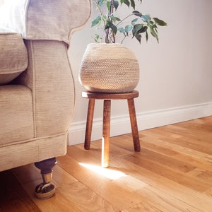 May include: A white woven plant basket containing a green leafy plant, resting on a wooden stool with four legs. The stool is positioned in front of a white sofa with beige fabric.