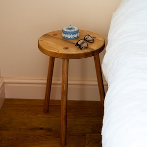 Può includere: Un tavolino rotondo in legno con tre gambe. Un contenitore decorativo blu e bianco e un paio di occhiali sono appoggiati sulla superficie del tavolo. Il tavolo è accanto a un letto bianco.