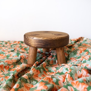 May include: A small, round wooden stool with four legs. The stool is made of dark brown wood and has a natural finish. It is sitting on a green and orange floral fabric.