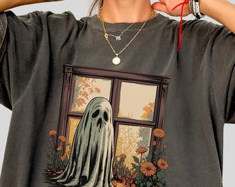 May include: Dark gray t-shirt featuring a ghost illustration. The ghost is depicted in a window with a fall scene, surrounded by flowers and leaves. The shirt has short sleeves and a relaxed fit, suitable for casual wear.