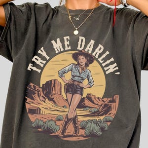 Pu&ograve; includere: T-shirt grigio scuro con un'illustrazione di un paesaggio desertico e una donna con cappello da cowboy. Il testo "TRY ME DARLIN'" &egrave; sopra l'immagine. La maglietta ha un aspetto vintage sbiadito.