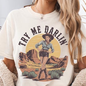 Pu&ograve; includere: T-shirt color avorio con grafica di una donna con cappello e stivali da cowboy, ambientata in un paesaggio desertico con la scritta "TRY ME DARLIN'". La maglietta ha una vestibilit&agrave; ampia.