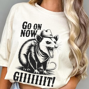 Może przedstawiać: Kremowa koszulka z czarno-białą ilustracją oposa w kapeluszu i chuście, z napisem "Go On Now GIIIIIIIT!". Koszulka jest w stylu casual unisex.