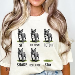 Op de afbeelding: Cr&egrave;mekleurig T-shirt met illustraties van een Franse Bulldog die commando's uitvoert: zit, ga liggen, apporteer, schud, rol om en blijf. De tekst voor elk commando staat onder de bijbehorende afbeelding.
