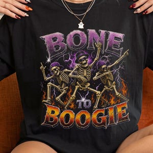 May include: A black t-shirt featuring a graphic of three skeletons dancing with purple lightning and orange flames in the background. The text "BONE TO BOOGIE" is printed in large, stylized letters. The skeletons wear hats and sunglasses.