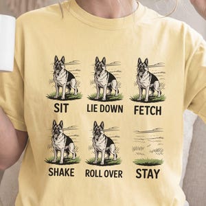 以下が含まれることがあります： バター色のTシャツ。ジャーマンシェパード犬が6つのポーズをとるグラフィックがプリントされています。各ポーズには太字で「sit, lie down, fetch, shake, roll over, stay」と書かれています。ゆったりとしたフィット感のTシャツです。