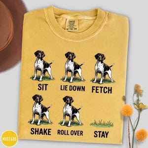 Può includere: T-shirt Comfort Colors color senape con sei illustrazioni di un cane bianco e nero che dimostra comandi: siediti, sdraiati, prendi, scuoti, rotola e resta. Il testo "SIT", "LIE DOWN", "FETCH", "SHAKE", "ROLL OVER" e "STAY" è stampato.