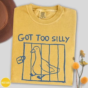 Op de afbeelding: Mosterdgeel T-shirt met de blauwe tekst "GOT TOO SILLY". Een blauwe illustratie van een eend in een kooi staat onder de tekst. Het shirt heeft een ronde hals en een Comfort Colors label.