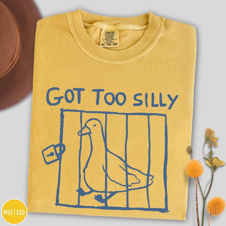 Op de afbeelding: Mosterdgeel T-shirt met de blauwe tekst "GOT TOO SILLY". Een blauwe illustratie van een eend in een kooi staat onder de tekst. Het shirt heeft een ronde hals en een Comfort Colors label.