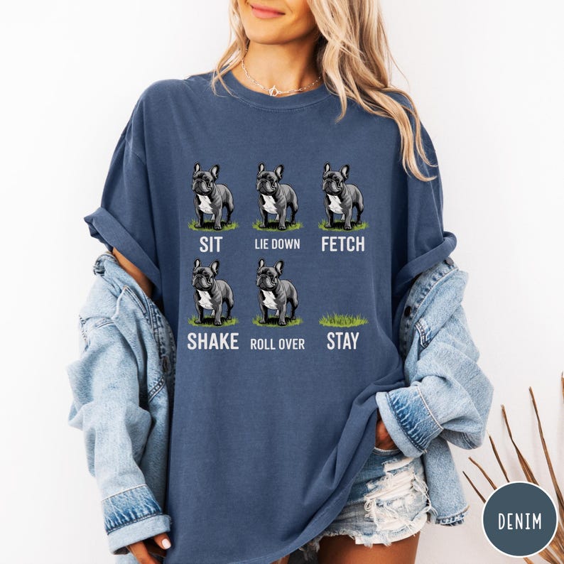 Op de afbeelding: Een marineblauw t-shirt met een afbeelding van zes Franse Bulldogs die commando's illustreren zoals "Sit", "Lie Down" en "Fetch". Het shirt toont ook de commando's "Shake", "Roll Over" en "Stay". Het woord "DENIM" staat rechtsonder.