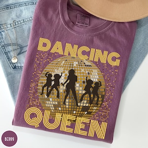 May include: Berry-colored t-shirt with the words "DANCING QUEEN" in yellow, along with a disco ball graphic and dancing silhouettes. The shirt is a Comfort Colors brand, size XL-TG-EG.
