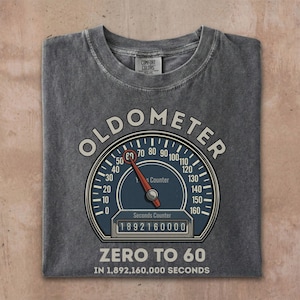 May include: Gray t-shirt with a graphic of a speedometer labeled "OLDOMETER." The speedometer needle points to 60, with the text "ZERO TO 60 IN 1,892,160,000 SECONDS" below. The shirt is made by Comfort Colors.