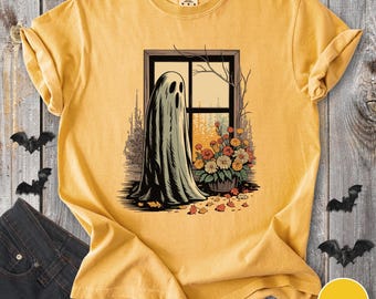 May include: Mustard-colored t-shirt featuring a graphic of a ghost standing by a window with flowers. The ghost is draped in a flowing sheet. The window reveals a fall scene. Bats are on the sides.