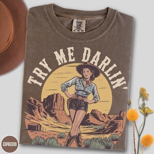 Pu&ograve; includere: T-shirt color espresso con grafica di una cowgirl in un paesaggio desertico. Il testo "TRY ME DARLIN'" &egrave; arcuato sopra la cowgirl. La maglietta &egrave; del marchio Comfort Colors, taglia XL-TG-EG.