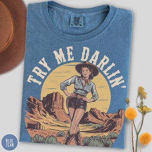 Pu&ograve; includere: T-shirt blu jeans Comfort Colors con un'illustrazione retr&ograve; di una cowgirl e la scritta "TRY ME DARLIN'". Il design include un paesaggio desertico con un sole al tramonto. La maglietta ha girocollo e maniche corte.