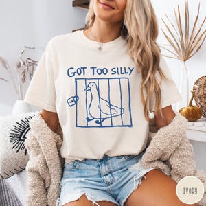 Op de afbeelding: Ivoor kleurig T-shirt met de tekst "GOT TOO SILLY" boven een blauwe illustratie van een vogel in een kooi. Het T-shirt heeft een relaxte pasvorm en korte mouwen. Het shirt is gemaakt van een zacht materiaal.
