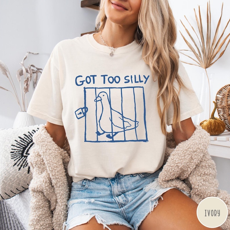 Op de afbeelding: Ivoor kleurig T-shirt met de tekst "GOT TOO SILLY" boven een blauwe illustratie van een vogel in een kooi. Het T-shirt heeft een relaxte pasvorm en korte mouwen. Het shirt is gemaakt van een zacht materiaal.