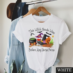 May include: White t-shirt with a graphic of a witch's hat, cauldron, wine glass, and a book with the text "This Witch Needs Wine Before Any Hocus Pocus".