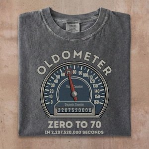 May include: Dark gray t-shirt with a graphic that reads "OLDOMETER" above a speedometer design. The speedometer needle points to 70, with the text "ZERO TO 70 IN 2,207,520,000 SECONDS" below. The shirt is made by Comfort Colors.